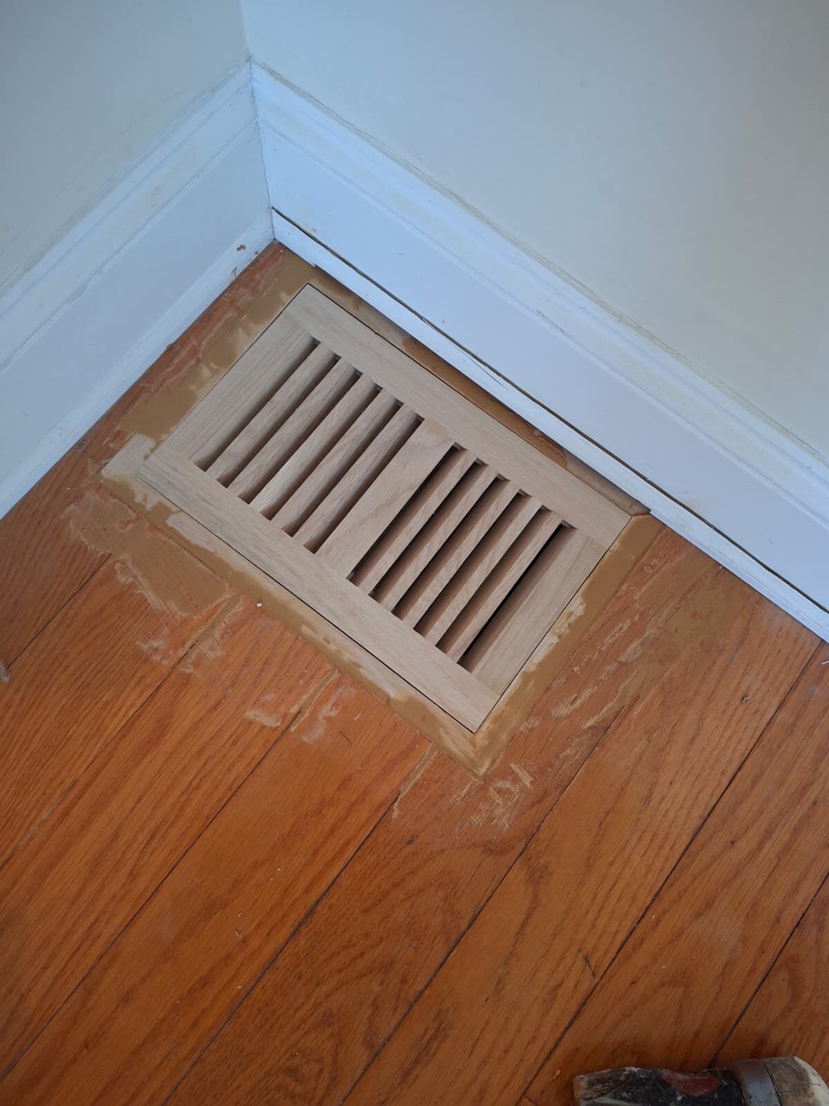 New oak floor vent grille installed flush in hardwood flooring at baseboard corner